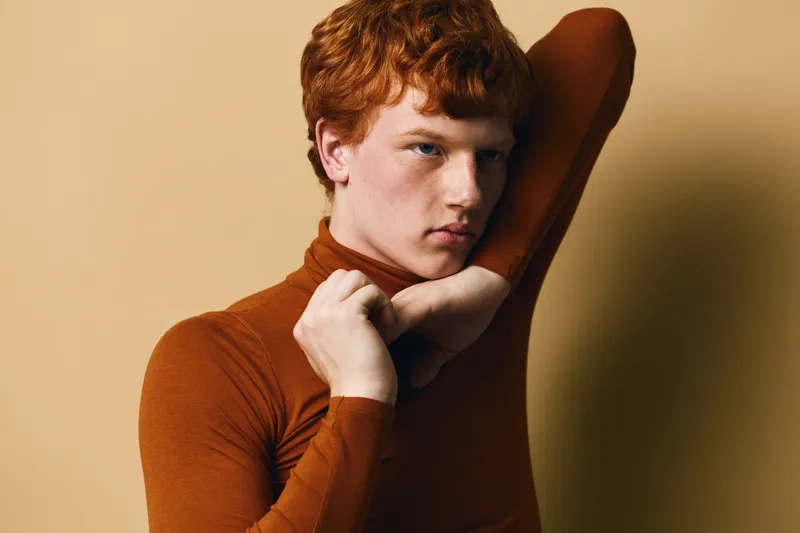 Portrait of young man with red hair wearing brown turtleneck, posing with hand near neck against beige background, showing serious expression and natural skin texture.