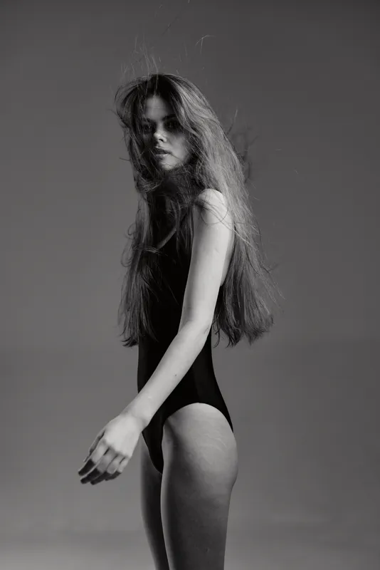 Black and white portrait of a young woman with long hair in a bodysuit, striking a confident pose against a plain background.