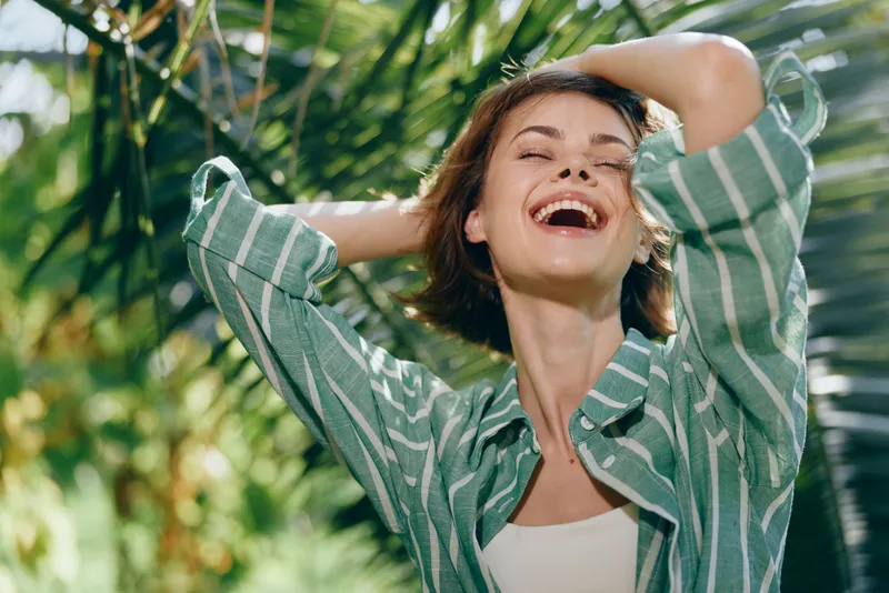 Woman outdoors, smile, a joyful woman stands with arms raised under bright sun, enjoying a carefree moment among lush greenery, radiating happiness and vitality.