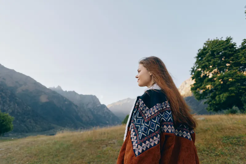 Woman portrait in sweater with long hair standing in mountain landscape meadow, looking sideways at distant peaks. Nature travel scene with trees, calm light, warm autumn mood and solitude.