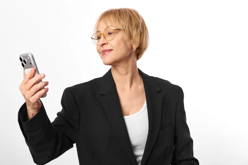 Senior woman in business suit and glasses holds a smartphone in studio portrait, looking at the screen with focused thinking, modern communication and technology