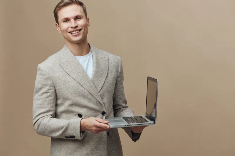 smiling young caucasian man holding laptop wearing beige jacket standing isolated on beige background remote work digital nomad technology concept