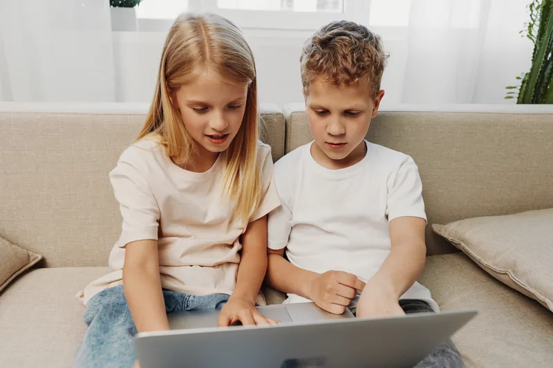 Two focused children sitting on couch using laptop together in bright room. Concept for education, technology, learning, childhood and digital interaction at home environment.