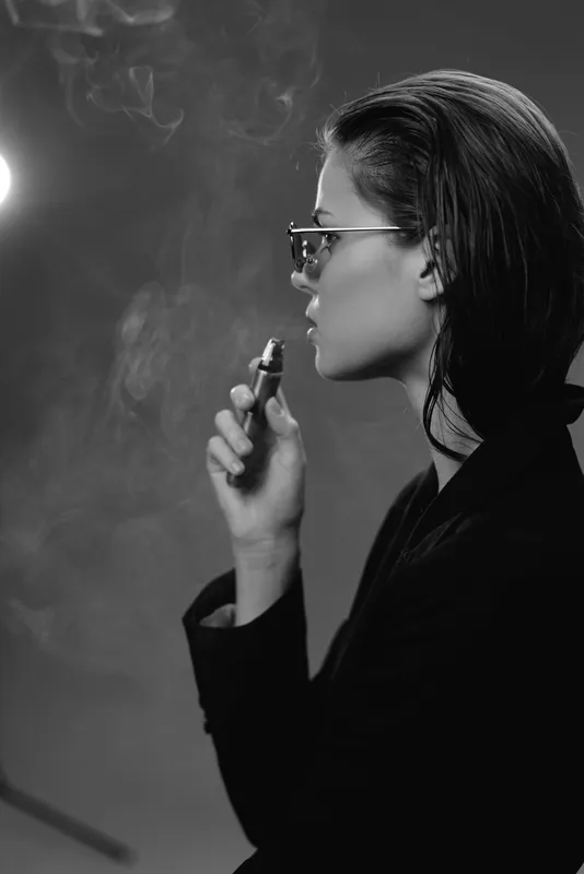 Black and white portrait of a woman vaping with glasses in a dark setting and smoke around her.