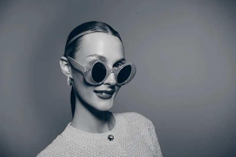 Smiling young Caucasian woman with pale skin wearing stylish round sunglasses and sweater in monochrome studio portrait. Concept of fashion, style, and confidence.