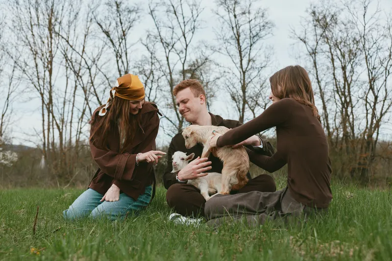 Three friends sit on grass in a park, sharing a joyful moment with a small dog, as they pet and play, showcasing casual outdoor friendship and warm connection in natural daylight