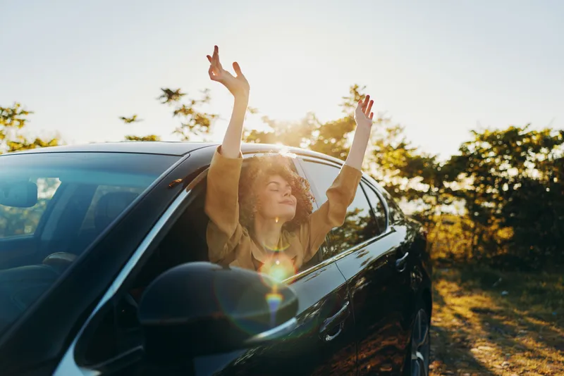 Joyful woman in a car, sunlit outdoors, arms raised, retro film color vibe, carefree moment, warm glow, open window, natural scenery, candid lifestyle