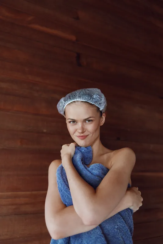 young woman wrapped in blue towel wearing shower cap smiling indoors with wooden background, natural light and relaxed mood, wellness and self-care concept