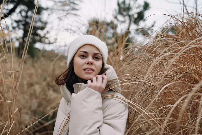 Outdoor portrait of a woman in a beige winter coat and knit hat among tall grasses, showcasing casual fashion, calm mood, and natural seasonal scenery in soft light and breeze.