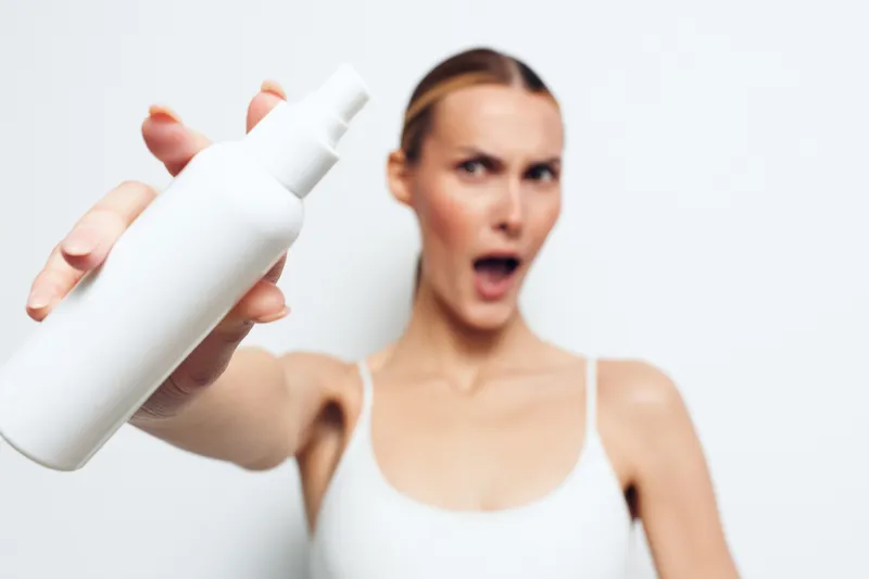 Woman skincare wellness spa beauty face spray bottle studio closeup, hand holds spray bottle near lens, surprised woman in white top behind, soft studio lighting on clean background