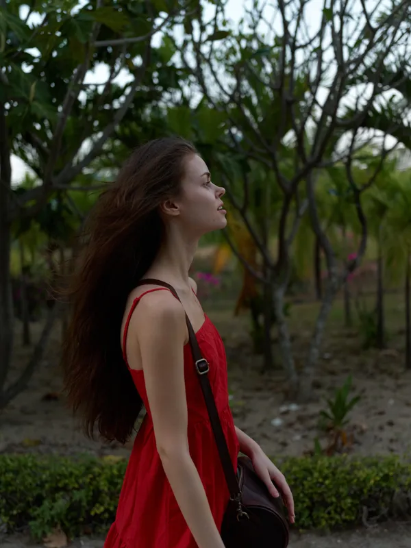 young woman in red dress standing outdoors among trees, looking up thoughtfully, natural light highlighting her profile and long hair, peaceful moment captured