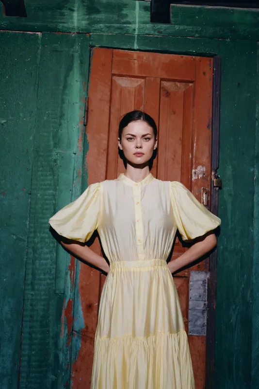 Stylish woman stands in a doorway wearing a flowing pale yellow dress posing with confidence, creating a striking portrait with bold fashion vibes and moody studio lighting.