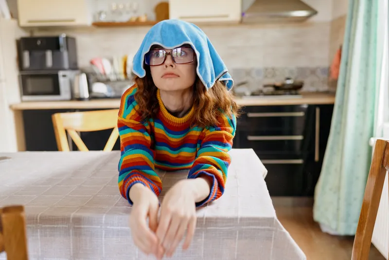 Cheerful child indoors in the kitchen, wearing a colorful striped sweater and a towel on the head, leaning forward with hands on a table, capturing a playful cozy home moment indoors now