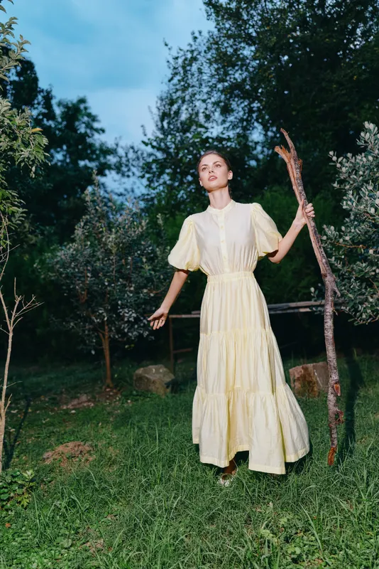 Woman wearing a yellow dress standing in a grassy garden outdoors holding a long stick, exploring a natural setting with trees and an open evening sky.