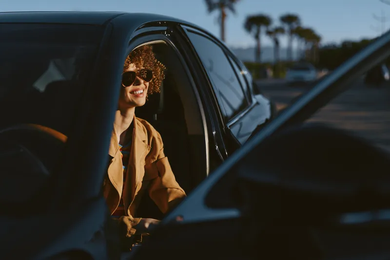 Smiling woman with curly hair inside a car wearing sunglasses, casual jacket, framed by warm retro colors. Close-up on a roadside scene, conveying lifestyle, travel, and self expression.