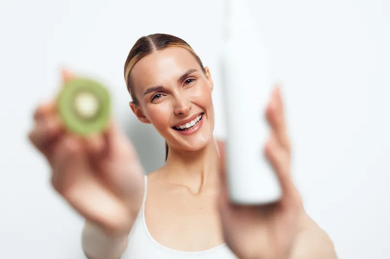 Smiling young woman holds fresh sliced kiwi and white bottle close to camera isolated on white background healthy lifestyle concept natural skincare and beauty product focus on face