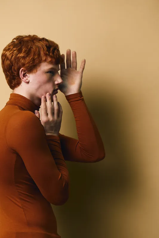 Young man with red hair wearing brown turtleneck sweater performs whisper gesture with hands near mouth against beige background, side view portrait of handsome male model.