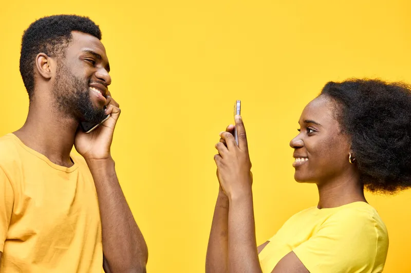 Happy young Black couple taking selfies with smartphones against a vibrant yellow background, showcasing joy and connection in modern lifestyle