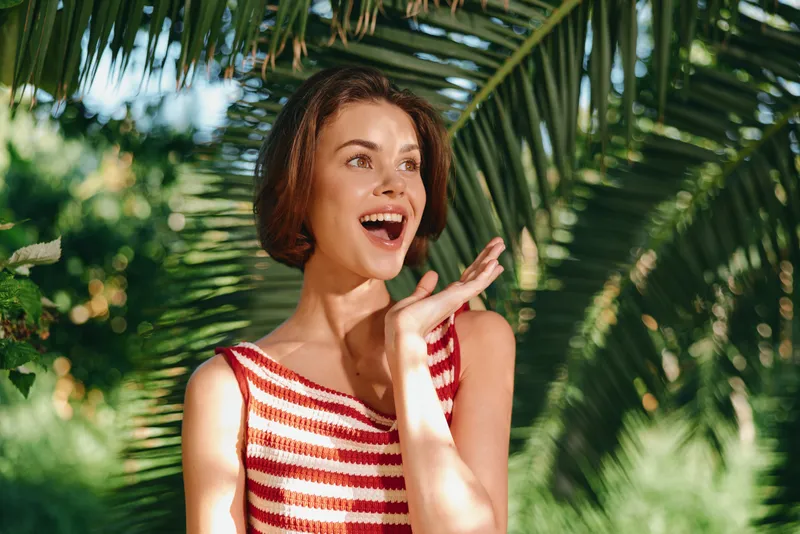 A cheerful woman in a striped sleeveless top stands outdoors among lush tropical plants, warmly lit by sunlight. She smiles brightly, radiating positive energy and relaxed confidence.