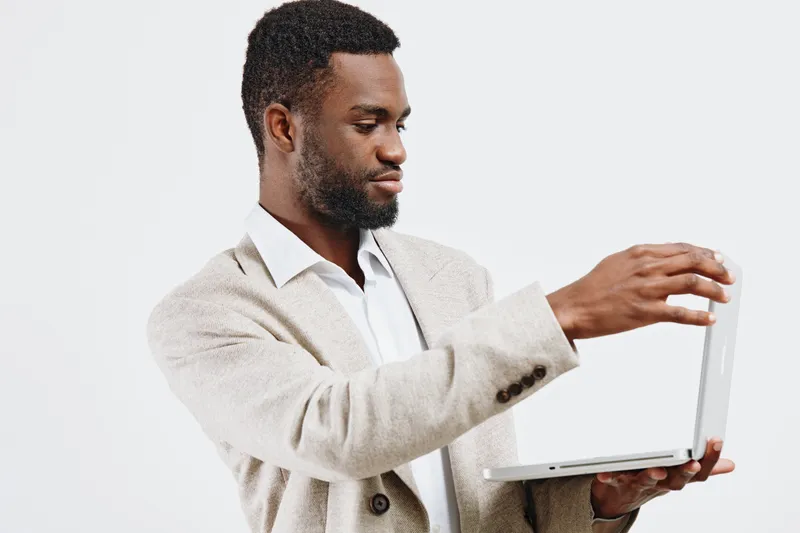Professional young man with short black hair and beard, wearing beige blazer and white shirt, holding open silver laptop in hand, looking at screen, neutral background, studio shot, business work conc