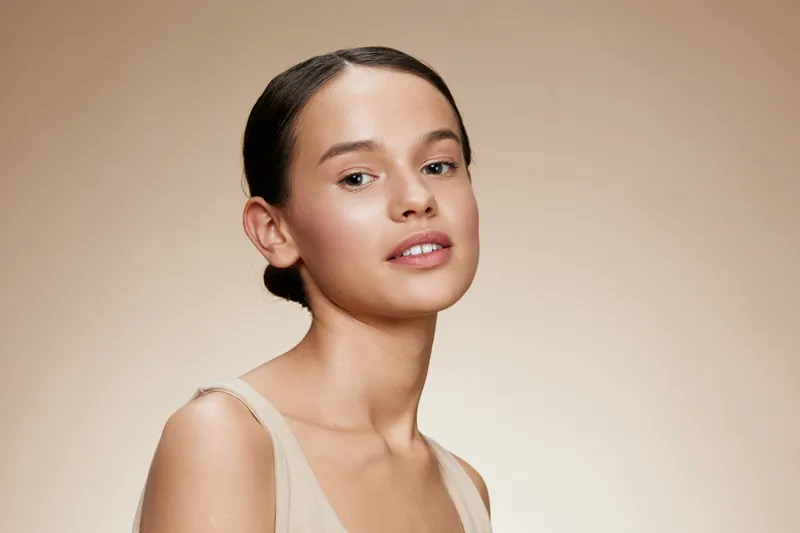 Young female model with a neutral expression, wearing a light, elegant outfit against a soft beige background, perfect for beauty and skincare concepts