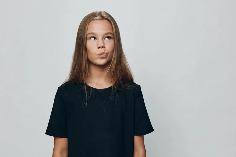 Teenage girl with long hair and black t shirt looking to side standing against gray background. Portrait of young female with thoughtful expression and casual style in studio.