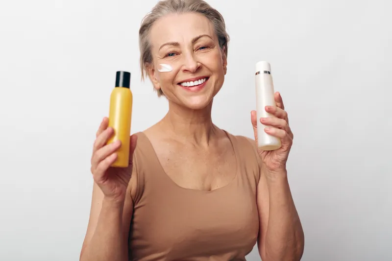 Smiling mature woman with short hair holds two skincare bottles and has cream on cheek. Concept of beauty routine, aging care, wellness, self-care, and natural skin health.