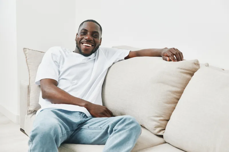 Happy young African man relaxing on a sofa, wearing casual white t shirt and jeans, expressing joy and comfort in a bright living room