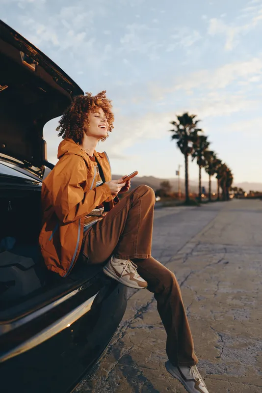 Woman lifestyle, sitting on car hood by urban road at sunset, curly haired woman in casual outfit leaning on vehicle, relaxed travel mood, warm evening light, outdoors, alone