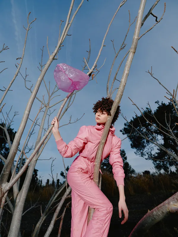 Fashion portrait of a model in a pink jumpsuit posing among bare trees with a pink balloon, capturing a whimsical outdoor scene with soft daylight and airy mood and subtle cinematic vibe