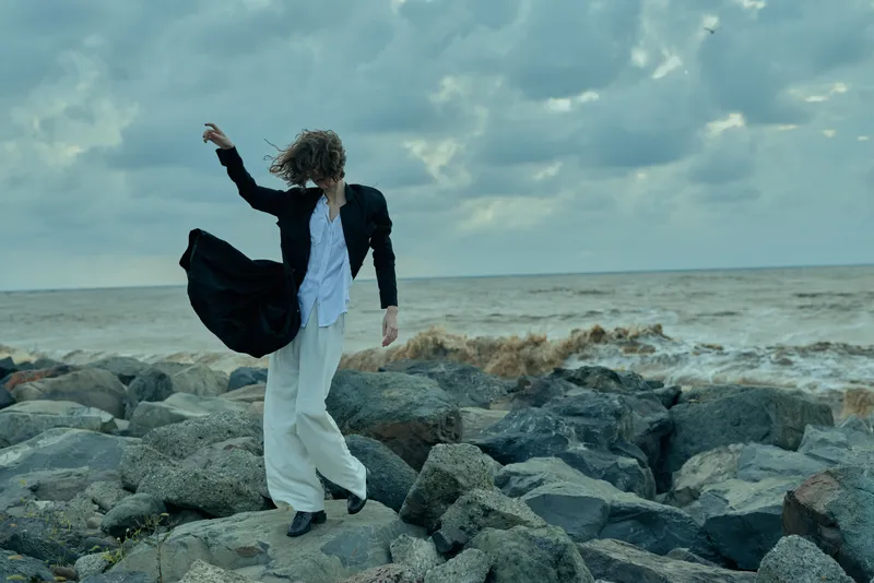 Model strolling along a windy rocky shore in a flowing white suit and dark jacket, dramatic sea backdrop, motion and fashion, outdoor photography for editorial use, capturing movement and weather