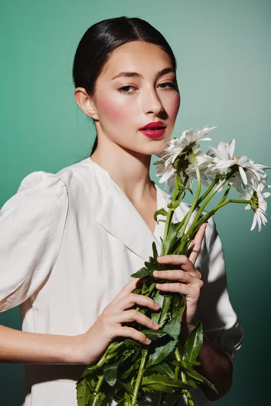 Young woman in white dress holds white daisies against green background in natural light. Beauty, creative fashion portrait with soft makeup and elegant hairstyle.