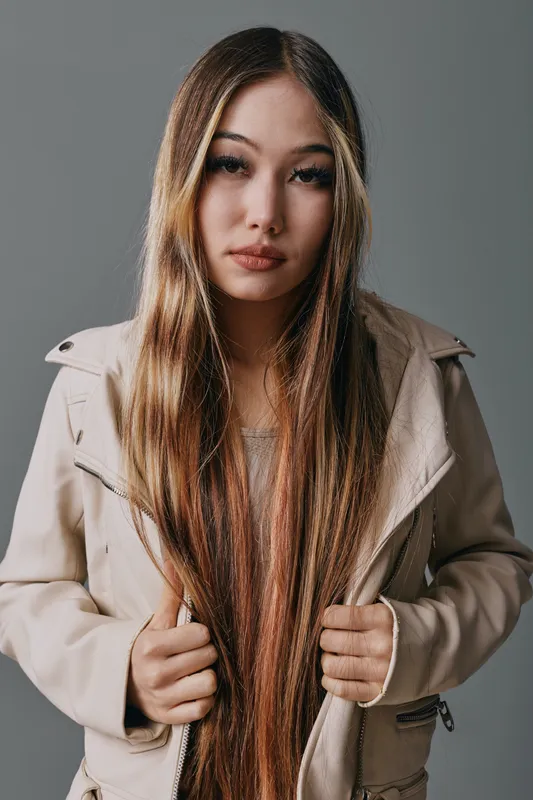 Young woman with long straight hair wearing beige jacket posing against gray background, natural makeup and serious expression for fashion portrait or hairstyle concept.
