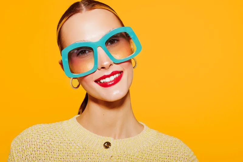 Smiling woman with bright red lipstick wearing large turquoise sunglasses and yellow sweater on vibrant orange background. Concept of fashion, summer style, and cheerful mood.