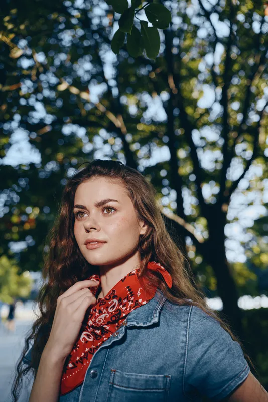 young woman wearing blue denim shirt and red scarf standing in sunlit park with green trees on a warm summer day