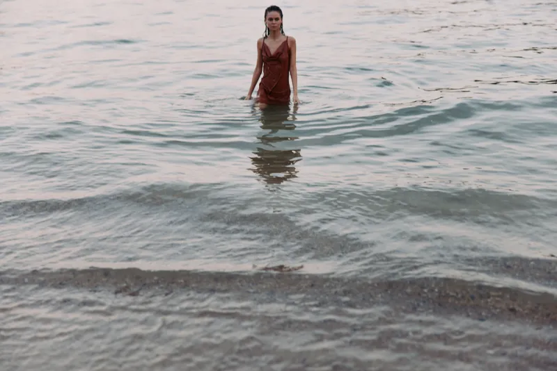 A solitary figure stands in shallow sea water near the shoreline, wearing a red swimsuit, with calm waves and soft sunlight creating tranquil tones.