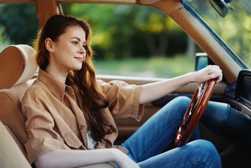 Young woman enjoying a peaceful drive in a vintage car, dressed in casual beige outfit, with long hair and a relaxed expression, set in a natural green background.