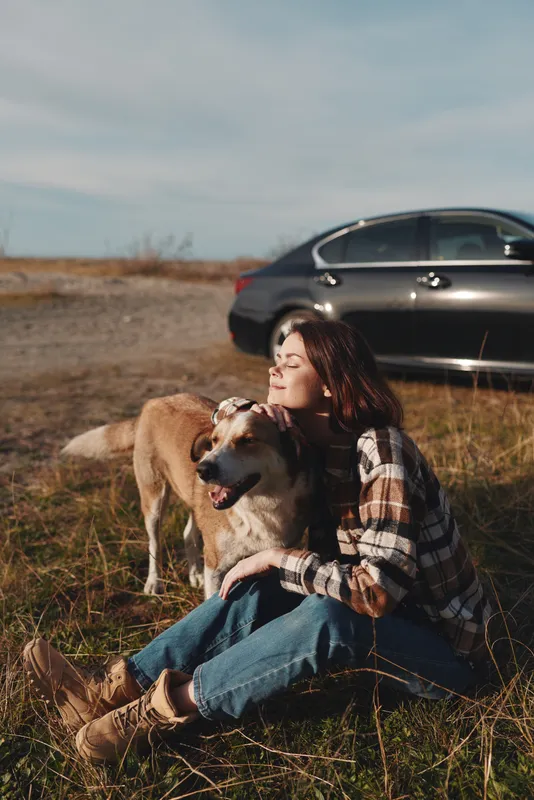 Woman sits on grass with her dog outdoors, leaning against a plaid shirt and jeans beside a parked car under a clear sky, sharing a calm moment of companionship in a rural setting with warm light