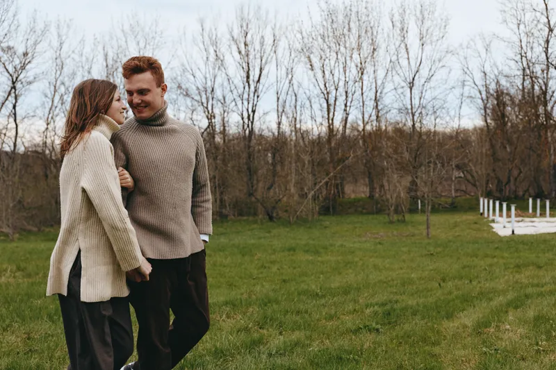 Couple outdoors, smiling and walking through a grassy field, cozy sweaters, natural light, gentle breeze, candid moment, soft colors, nature backdrop, relaxed lifestyle concept