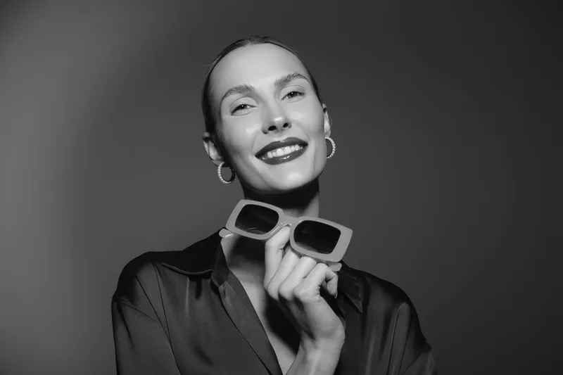 Happy white woman with light skin holding sunglasses smiles with closed eyes, wearing hoop earrings and dark clothes. Studio portrait in black and white, concept of fashion and joy.