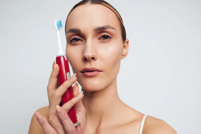 A young woman holds a red electric toothbrush near her face, looking directly at the camera with a neutral expression on a plain white background. Concept for dental care and hygiene.