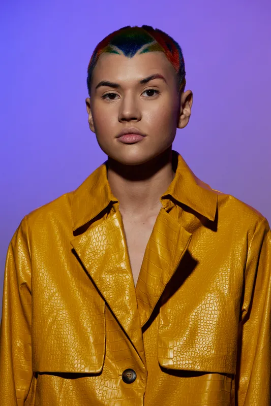 young woman with colorful short hair wearing a mustard yellow textured jacket standing against a purple background, expressing calm confidence and modern fashion style.