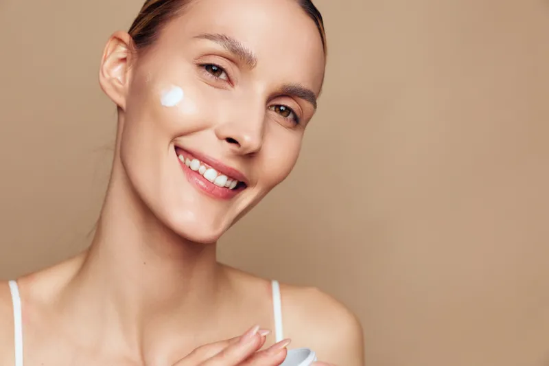 Smiling young woman applies cream on her cheek with natural makeup and clear skin isolated on beige background holding white jar of moisturizer for skincare routine and beauty care.