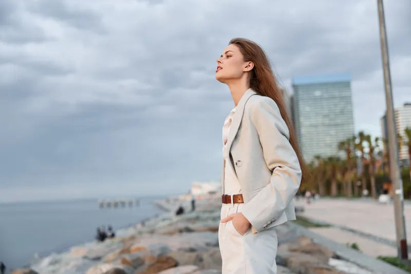 Confident woman in a business suit standing by the coast, exuding professionalism and determination with a modern skyline in the background.