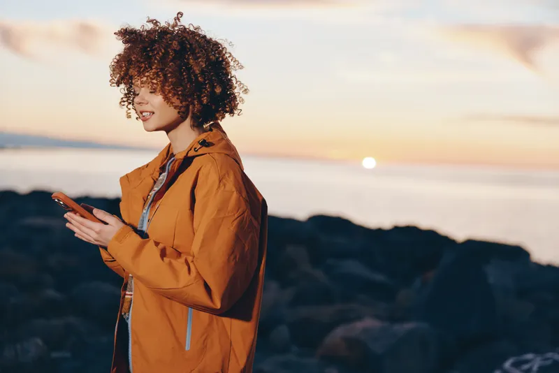 Woman with curly hair wearing an orange jacket stands at the seaside during sunset looking joyful and relaxed in an outdoor lifestyle moment soft coastal light by the shore today