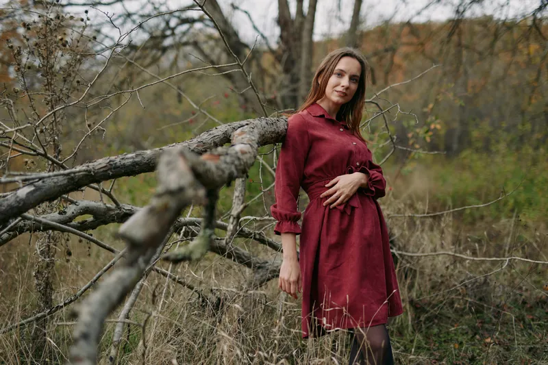 A candid portrait of a woman in a deep red dress leaning against a weathered branch, conveying authenticity and credibility in a quiet natural setting.