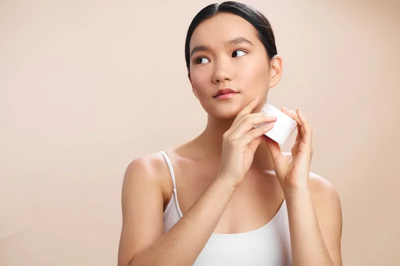 asian woman in a white tank top gently applies skincare in a bright, minimal space, exuding calm and self care while focusing on a subtle beauty routine and healthy daily habits