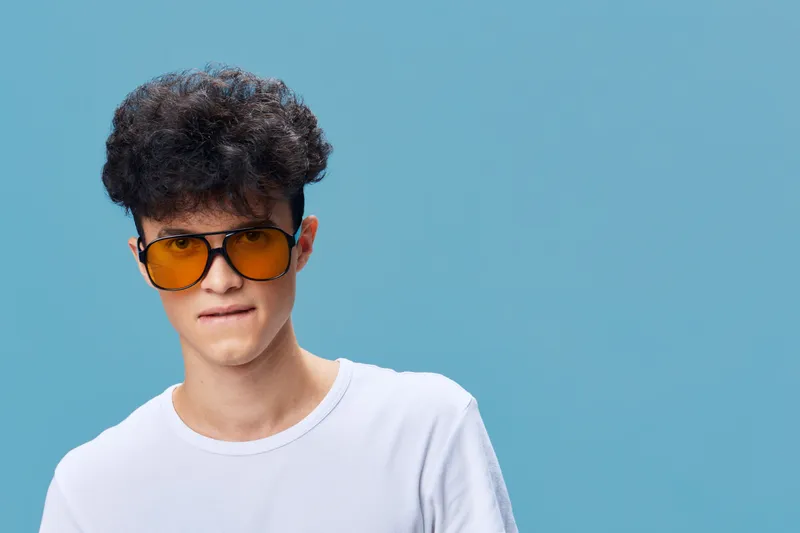 Young man with curly hair wearing large orange sunglasses and a white t-shirt, posing against a solid blue background with a playful expression.