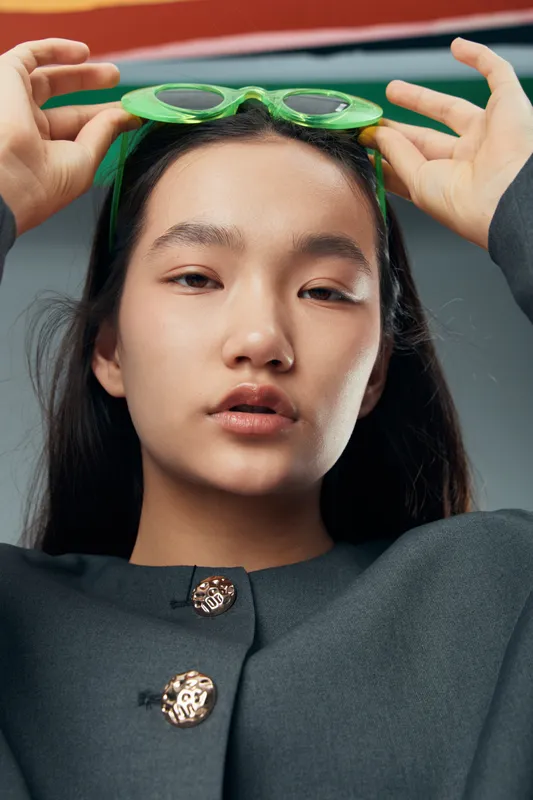 Young woman with straight black hair wearing dark gray coat and holding green sunglasses on head. Closeup portrait with neutral expression and smooth skin in studio light.