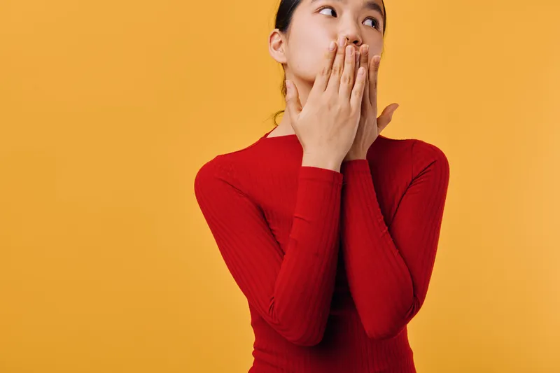 Woman in red sweater covering mouth with hands looking aside on yellow background for surprise, emotion, reaction, expression, and gesture concept of young adult female.